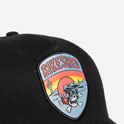 A black snapback baseball hat with a colorful patch on the front featuring a cartoonish motorcycle skull and bike shed logo - detail