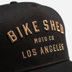A black trucker cap with 'BIKE SHED LOS ANGELES' embroidered in gold on the front.