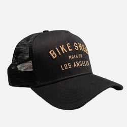 A black trucker cap with 'BIKE SHED LOS ANGELES' embroidered in gold on the front.