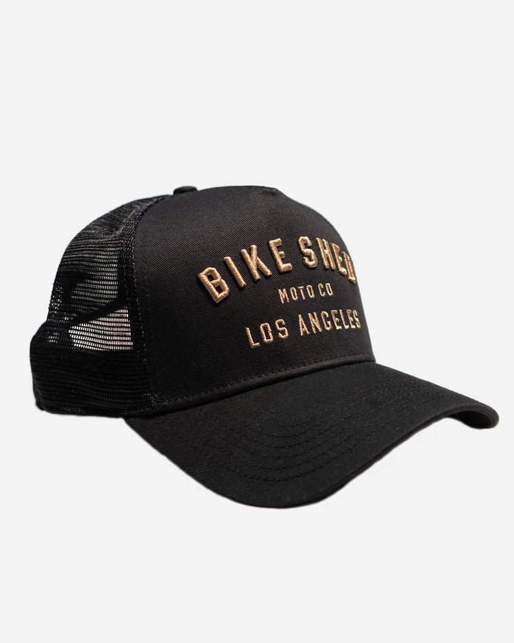 A black trucker cap with 'BIKE SHED LOS ANGELES' embroidered in gold on the front.