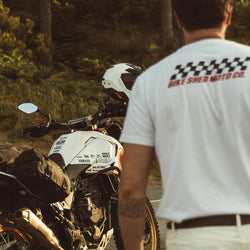 Model wearing Motorcycle Desert Race white t-shirt with a Bike Shed Moto logo, standing next to a motorcycle in a forest.