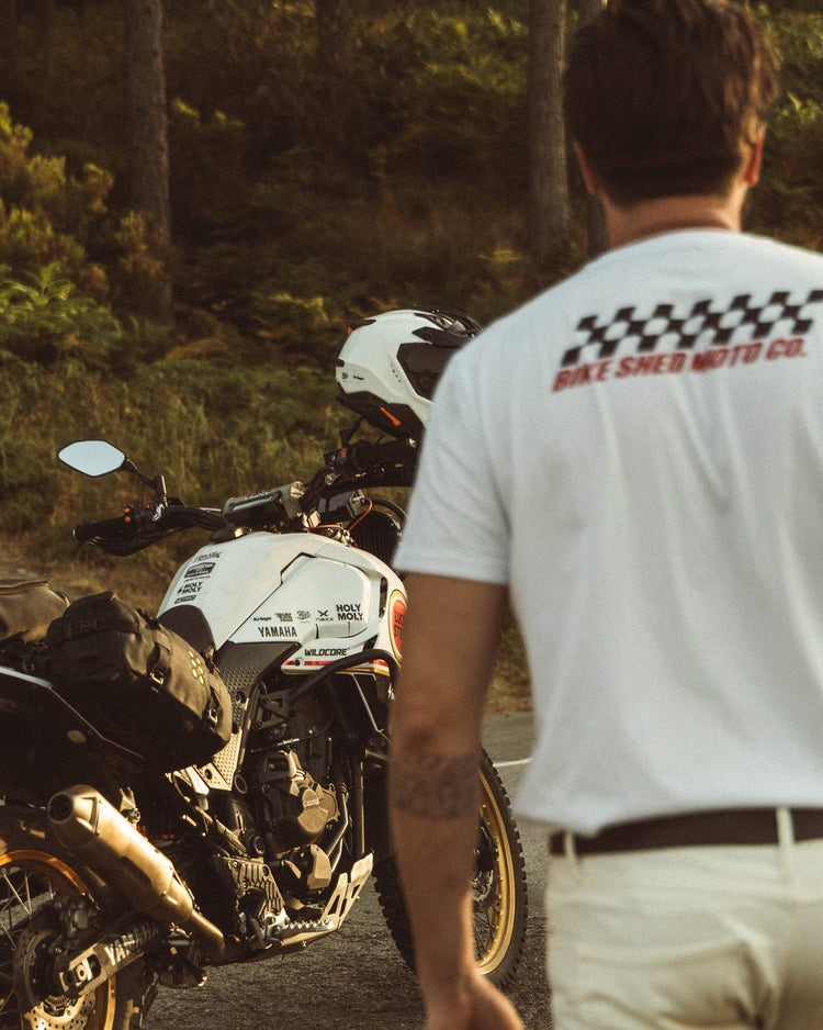 Model wearing Motorcycle Desert Race white t-shirt with a Bike Shed Moto logo, standing next to a motorcycle in a forest.