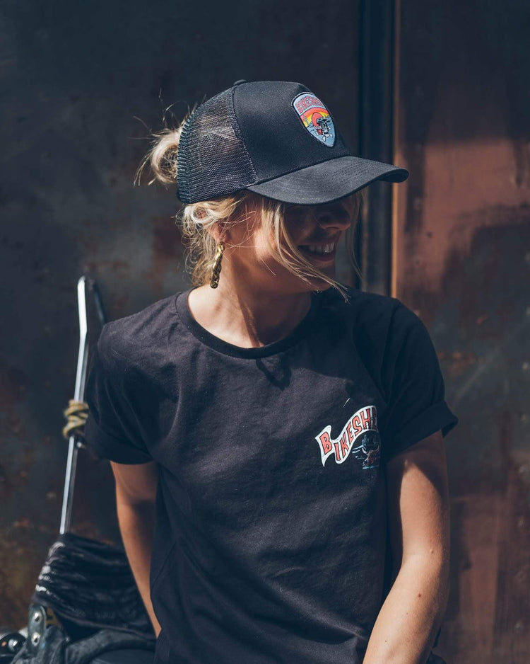 A black snapback baseball hat with a colorful patch on the front featuring a cartoonish motorcycle skull and bike shed logo - worn by a model