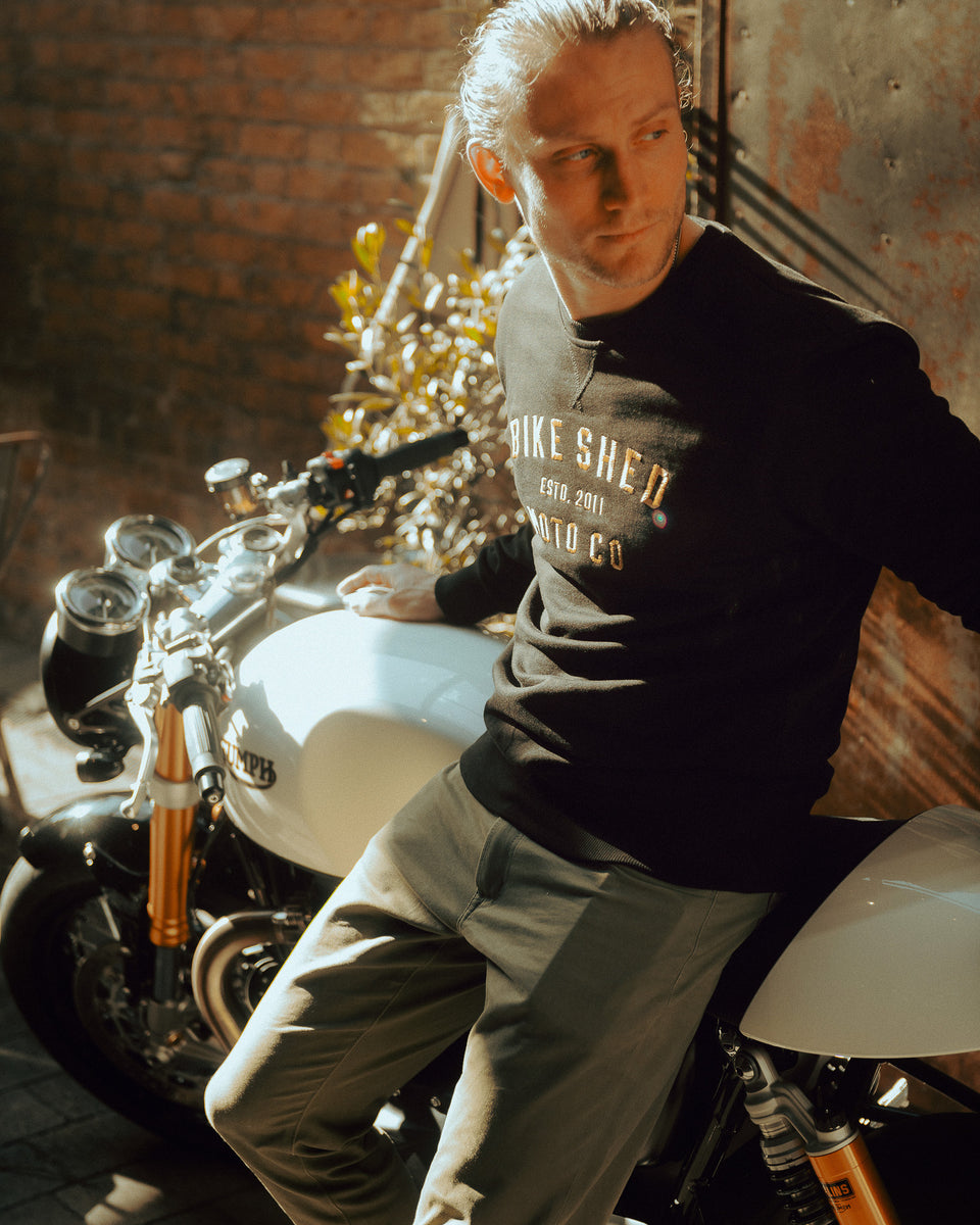 Model wearing black Bike Shed Moto Co logo Sweatshirt next to a motorcycle.