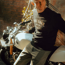 Model wearing black Bike Shed Moto Co logo Sweatshirt next to a motorcycle.