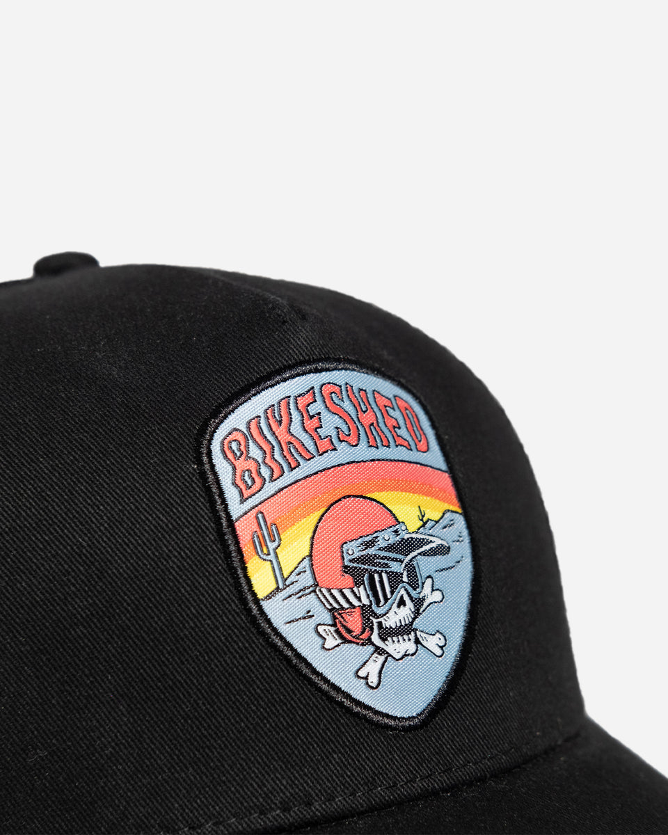 A black snapback baseball hat with a colorful patch on the front featuring a cartoonish motorcycle skull and bike shed logo - detail