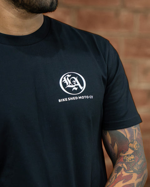 Model wearing a black Bike Shed Moto Co t-shirt with a logo against a brick wall