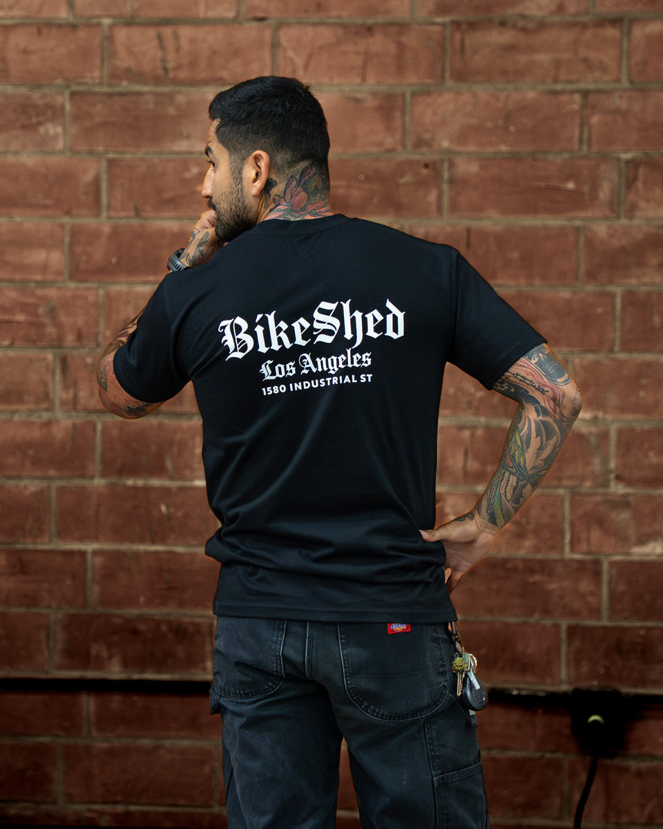 Model wearing a black Bike Shed Moto Co t-shirt with a logo against a brick wall