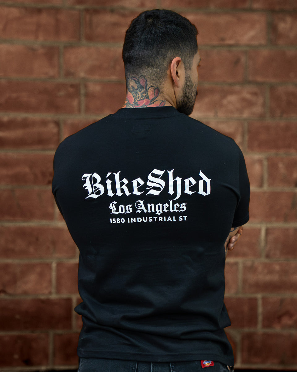 Model wearing a black Bike Shed Moto Co t-shirt with a logo against a brick wall