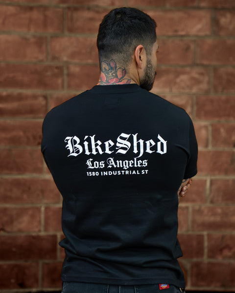 Model wearing a black Bike Shed Moto Co t-shirt with a logo against a brick wall