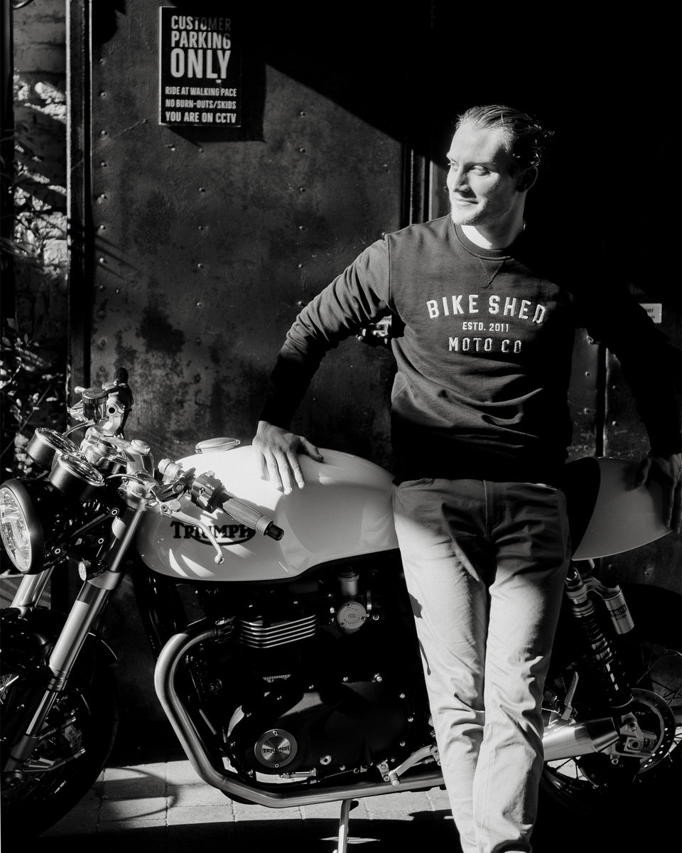 Model wearing black Bike Shed Moto Co logo Sweatshirt next to a motorcycle.