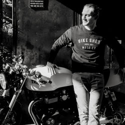 Model wearing black Bike Shed Moto Co logo Sweatshirt next to a motorcycle.