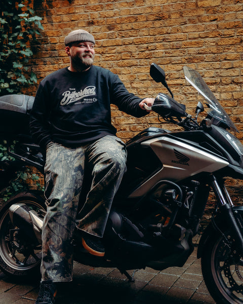 Model wearing Bike Shed Moto Co Black Applique Sweatshirt next to a motorcycle on a brick driveway