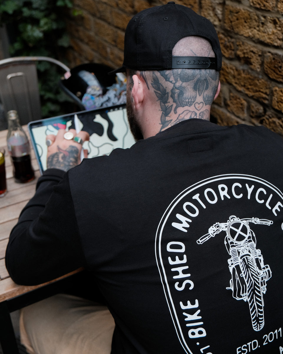 A long sleeve black T-shirt with a round neckline, featuring a front and back print. The front print is a logo, and the back print depicts an illustration of a motorcycle.