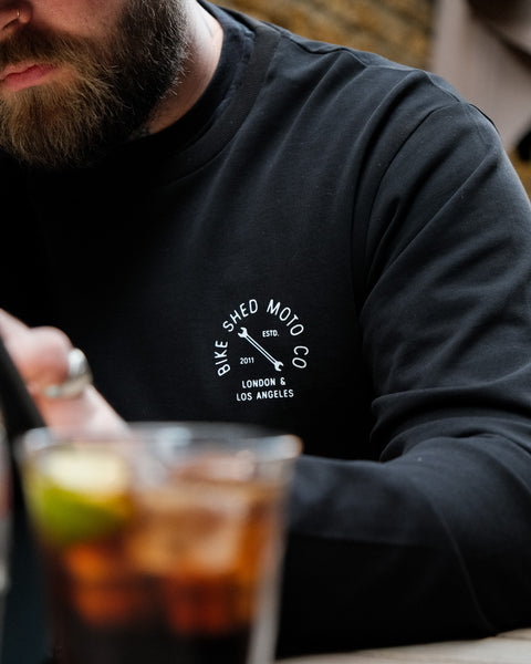 A long sleeve black T-shirt with a round neckline, featuring a front and back print. The front print is a logo, and the back print depicts an illustration of a motorcycle.