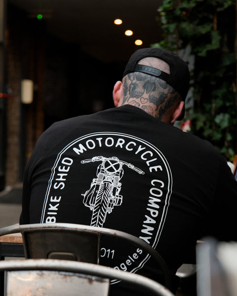 A long sleeve black T-shirt with a round neckline, featuring a front and back print. The front print is a logo, and the back print depicts an illustration of a motorcycle.