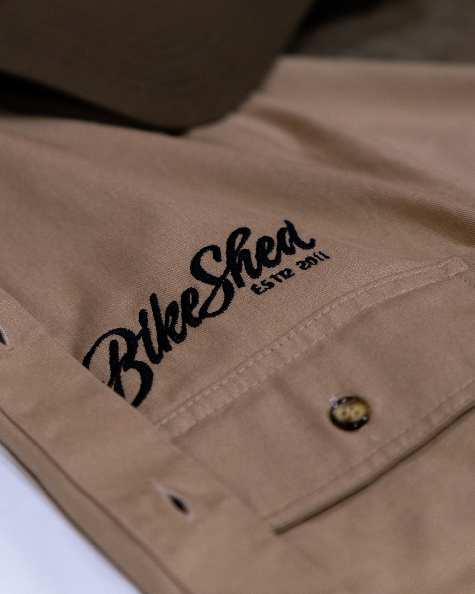 A beige Bike Shed Moto Co. Pennant shirt with button-up closure and chest pocket, featuring detailed embroidery on the chest and back.