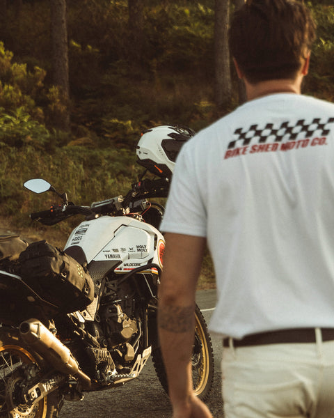 Model wearing Motorcycle Desert Race white t-shirt with a Bike Shed Moto logo, standing next to a motorcycle in a forest.