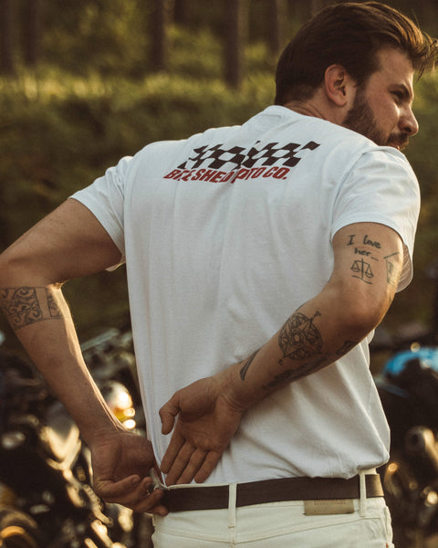 Model wearing Motorcycle Desert Race white t-shirt with a Bike Shed Moto logo, standing in a forest.