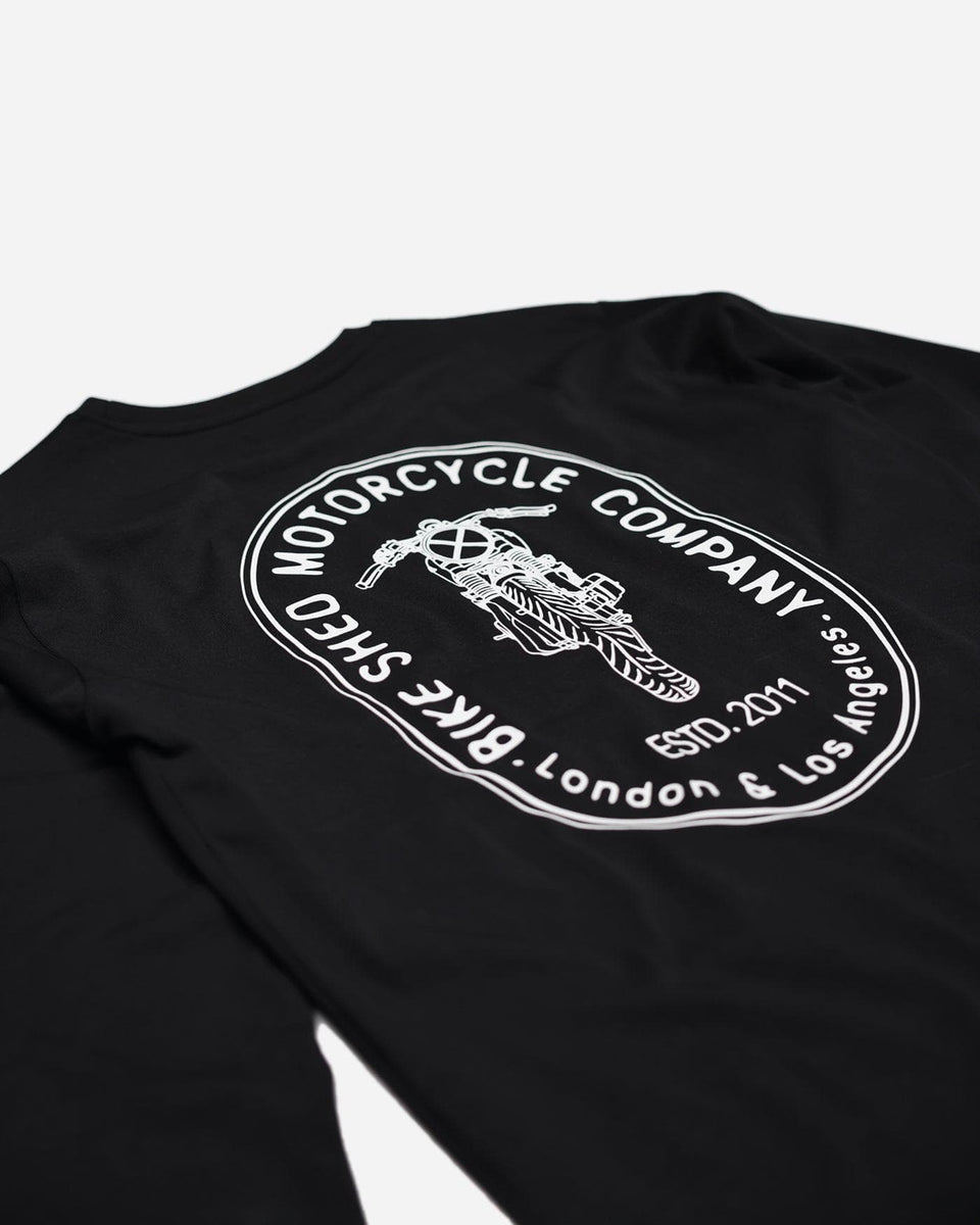 A long sleeve black T-shirt with a round neckline, featuring a front and back print. The front print is a logo, and the back print depicts an illustration of a motorcycle.