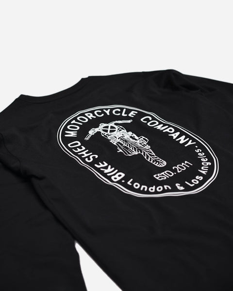 A long sleeve black T-shirt with a round neckline, featuring a front and back print. The front print is a logo, and the back print depicts an illustration of a motorcycle.