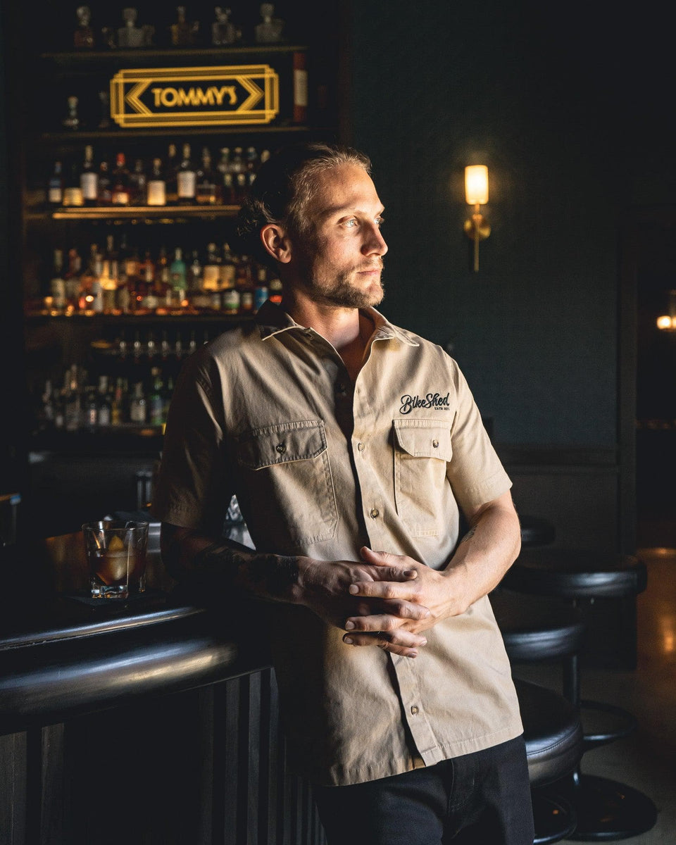 A beige Bike Shed Moto Co. Pennant shirt with button-up closure and chest pocket, featuring detailed embroidery on the chest and back.