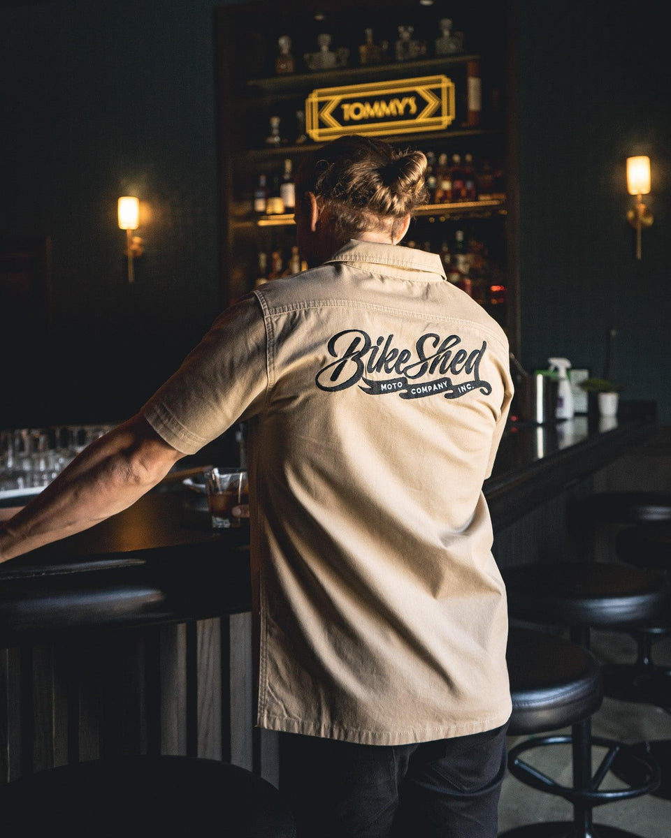 A beige Bike Shed Moto Co. Pennant shirt with button-up closure and chest pocket, featuring detailed embroidery on the chest and back.