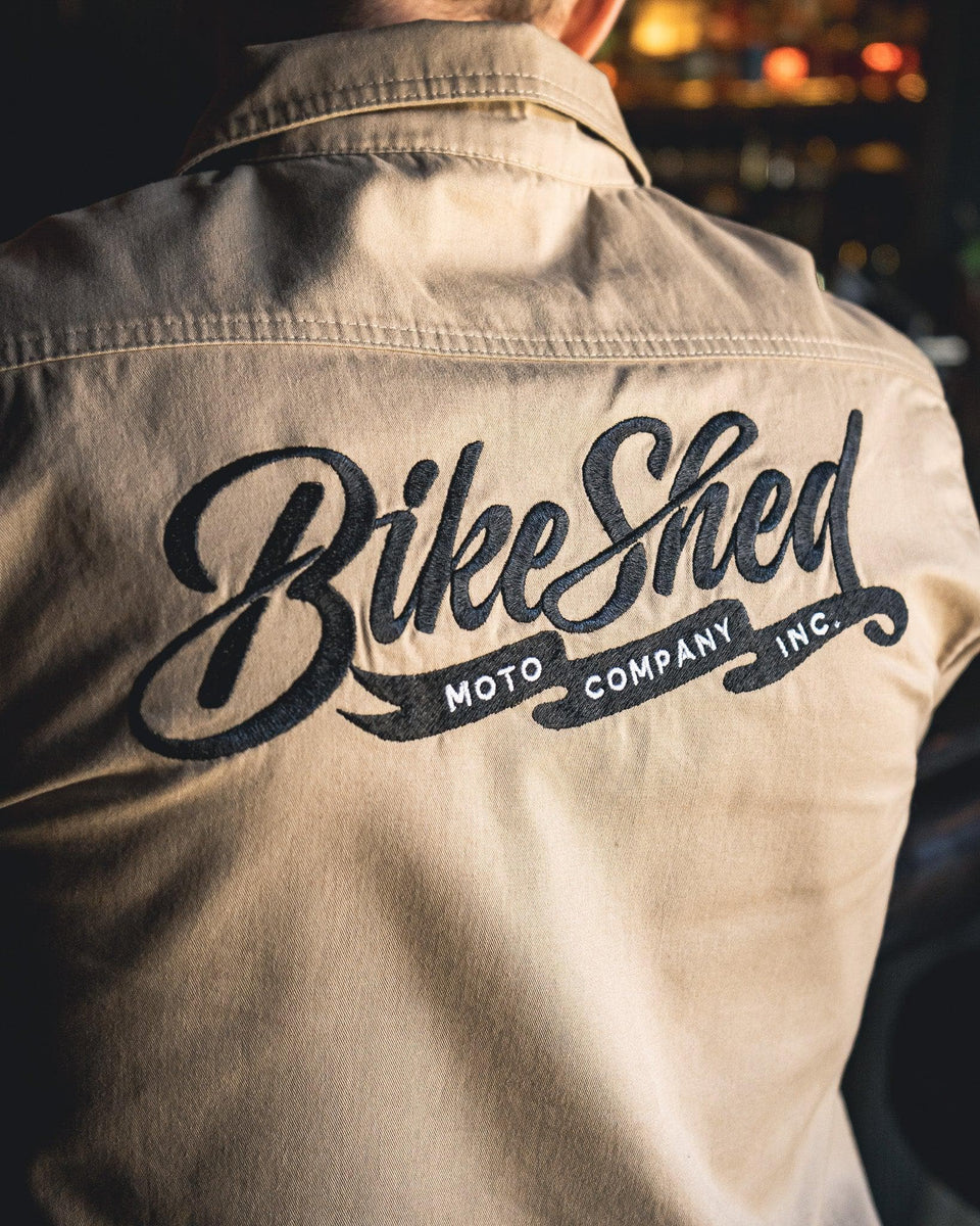 A beige Bike Shed Moto Co. Pennant shirt with button-up closure and chest pocket, featuring detailed embroidery on the chest and back.