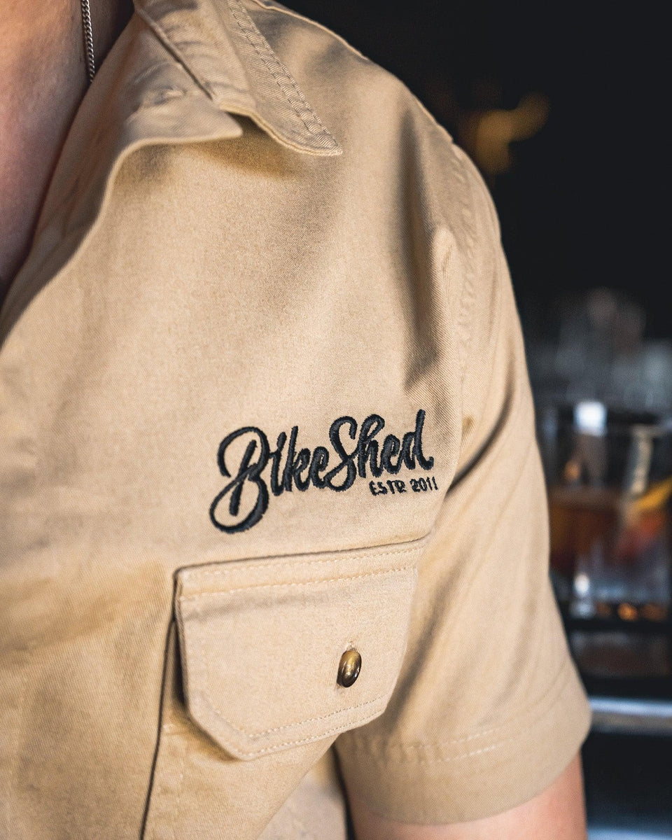 A beige Bike Shed Moto Co. Pennant shirt with button-up closure and chest pocket, featuring detailed embroidery on the chest and back.