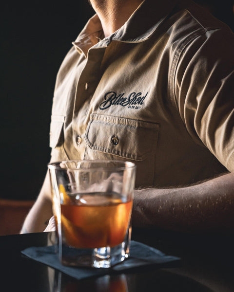 A beige Bike Shed Moto Co. Pennant shirt with button-up closure and chest pocket, featuring detailed embroidery on the chest and back.