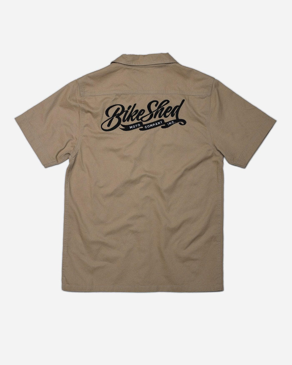 A beige Bike Shed Moto Co. Pennant shirt with button-up closure and chest pocket, featuring detailed embroidery on the chest and back.
