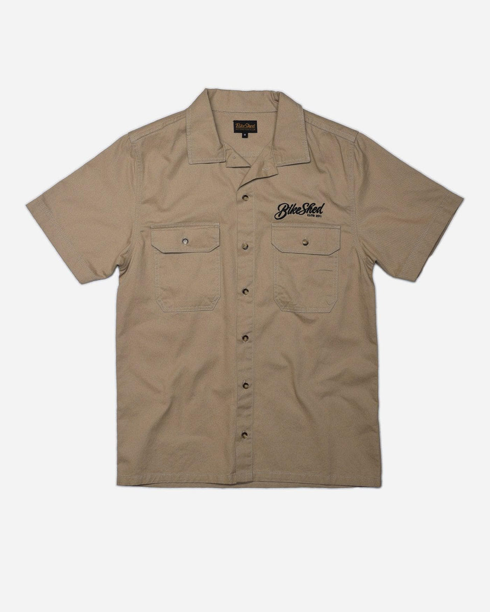 A beige Bike Shed Moto Co. Pennant shirt with button-up closure and chest pocket, featuring detailed embroidery on the chest and back.