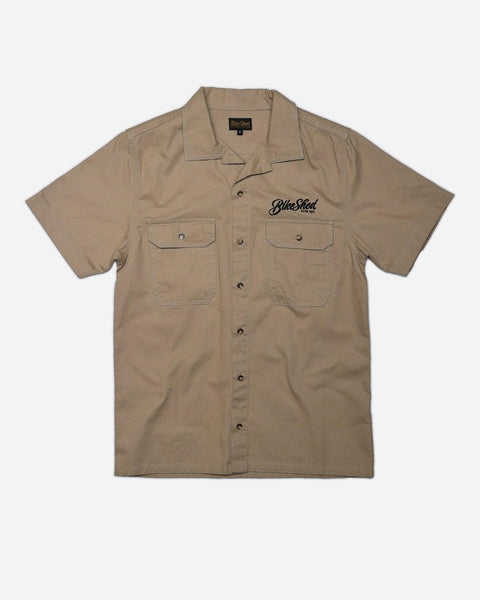 A beige Bike Shed Moto Co. Pennant shirt with button-up closure and chest pocket, featuring detailed embroidery on the chest and back.