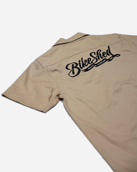 A beige Bike Shed Moto Co. Pennant shirt with button-up closure and chest pocket, featuring detailed embroidery on the chest and back.
