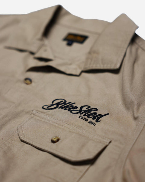 A beige Bike Shed Moto Co. Pennant shirt with button-up closure and chest pocket, featuring detailed embroidery on the chest and back.