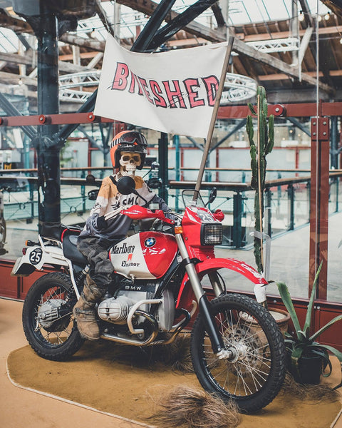Dead Dakar Doug sitting on a Dakar BMW R100 holding a Bike Shed flag